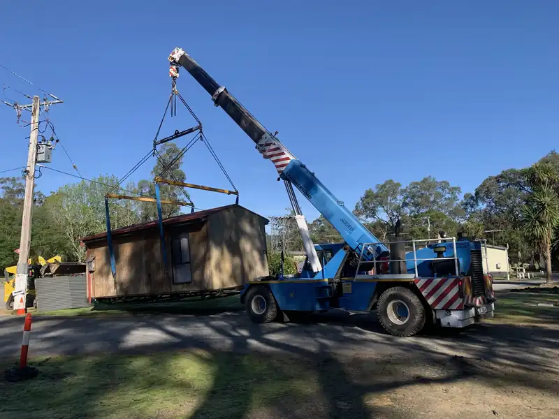 Blue crane lifting a timber cabin on a spreader frame beside a power pole